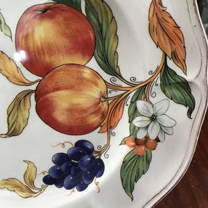 May include: A white ceramic plate with a hand-painted design of fruit and flowers. The design features red and yellow apples, blue grapes, orange berries, and white flowers.
