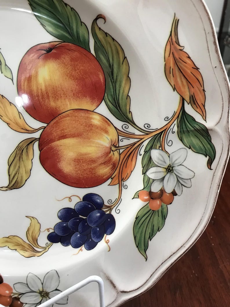 May include: A white ceramic plate with a hand-painted design of fruit and flowers. The design features red and yellow apples, blue grapes, orange berries, and white flowers.