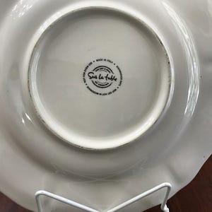 May include: White ceramic plate with a scalloped edge. The bottom of the plate has a black and white logo that reads "Sur La Table The Art & Soul of Cooking" and "May Get Hot in Microwave Not for Oven Use Made in Italy Dishwasher Safe".