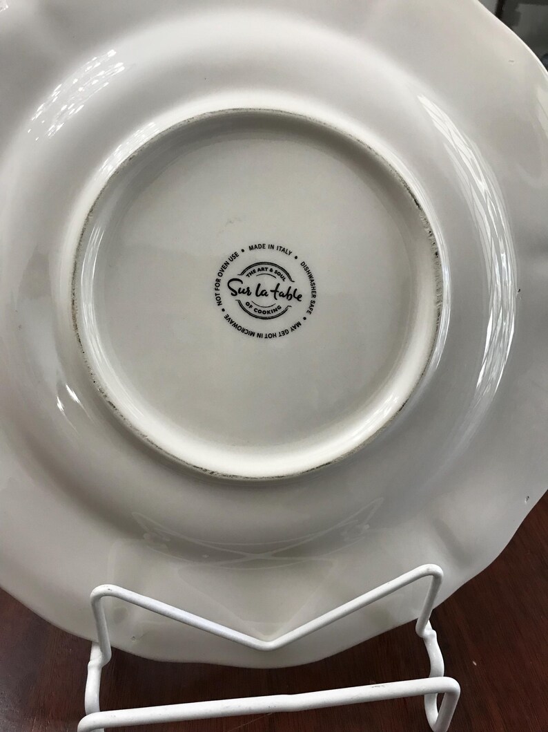 May include: White ceramic plate with a scalloped edge. The bottom of the plate has a black and white logo that reads "Sur La Table The Art & Soul of Cooking" and "May Get Hot in Microwave Not for Oven Use Made in Italy Dishwasher Safe".