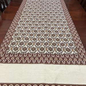 May include: A white table runner with a brown and gold paisley pattern and a red and gold border. The runner has a fringe edge.