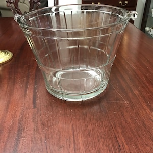 May include: A clear glass ice bucket with a flared shape and vertical ribbed detailing. The bucket has two small handles on either side of the rim. The bucket is sitting on a wooden table.