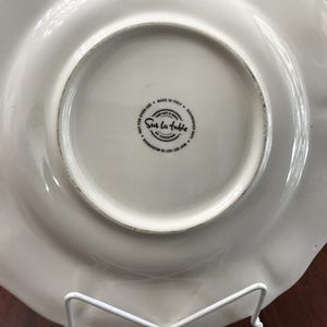 May include: White ceramic dinner plate with a scalloped edge. The plate has a black and white logo that reads "Sur La Table The Art & Soul of Cooking" and "Made in Italy, Dishwasher Safe, May Get Hot in Microwave, Not for Oven Use".
