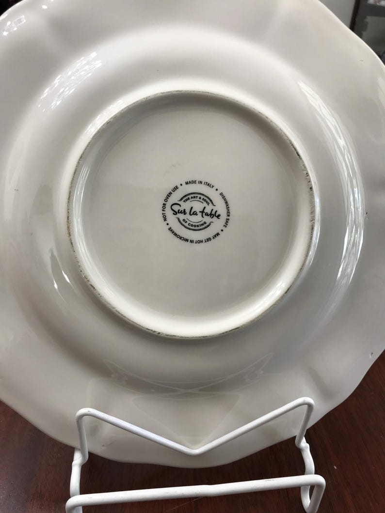 May include: White ceramic dinner plate with a scalloped edge. The plate has a black and white logo that reads "Sur La Table The Art & Soul of Cooking" and "Made in Italy, Dishwasher Safe, May Get Hot in Microwave, Not for Oven Use".