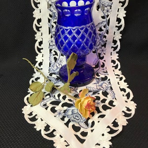 May include: A cobalt blue glass vase with a cut glass design sits on a white lace table runner. A single dried rose with a yellow center is next to the vase.