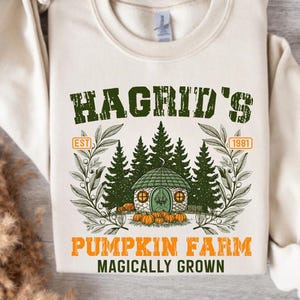 Puede incluir: Sudadera color crema con un gráfico verde y naranja. El gráfico dice "Hagrid's Pumpkin Farm" con una ilustración de una pequeña casa, árboles y calabazas. El texto "Magically Grown" está debajo.