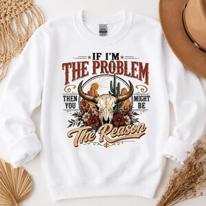 Può includere: Una felpa bianca a maniche lunghe con una stampa grafica raffigurante un teschio di bue, un cactus e formazioni rocciose desertiche. Il testo recita: IF I'M THE PROBLEM THEN YOU MIGHT BE THE REASON. Il design include fiori rossi e bianchi e fogliame.