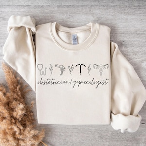Könnte beinhalten: Cremefarbenes Sweatshirt mit schwarzen Strichzeichnungen von medizinischen Instrumenten und floralen Elementen. Der Text "obstetrician/gynecologist" ist darunter aufgedruckt. Das Sweatshirt hat lange Ärmel und einen Rundhalsausschnitt.