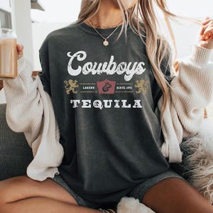 Pu&ograve; includere: T-shirt grigio scuro con la scritta "Cowboys Tequila" in lettere bianche e dorate. La maglietta presenta un design vintage con uno stemma e le parole "Legend & Since 1873".