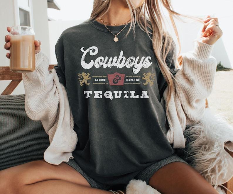 Pu&ograve; includere: T-shirt grigio scuro con la scritta "Cowboys Tequila" in lettere bianche e dorate. La maglietta presenta un design vintage con uno stemma e le parole "Legend & Since 1873".