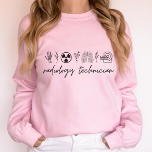 May include: A light pink sweatshirt with the words "radiology technician" in a cursive font. Above the text are black line drawings of a hand, a radiation symbol, flowers, lungs, and a CT scanner.