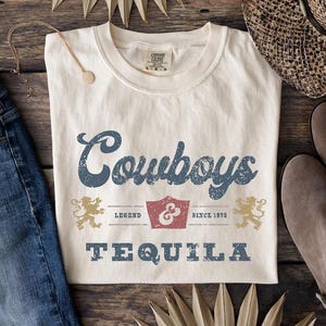 Pu&ograve; includere: T-shirt color crema con la scritta "Cowboys TEQUILA" in un carattere blu invecchiato. La maglietta presenta una grafica a scudo e due emblemi di leone. La maglietta &egrave; abbinata a jeans blu, occhiali da sole e stivali.