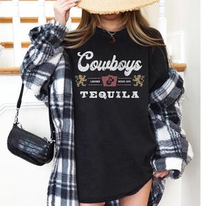 Pu&ograve; includere: T-shirt nero girocollo con le parole "Cowboys & Tequila" in stile vintage. La maglietta ha una grafica a scudo e la scritta "Legend Since 1895". La maglietta &egrave; indossata con una giacca a quadri e una borsa nera.
