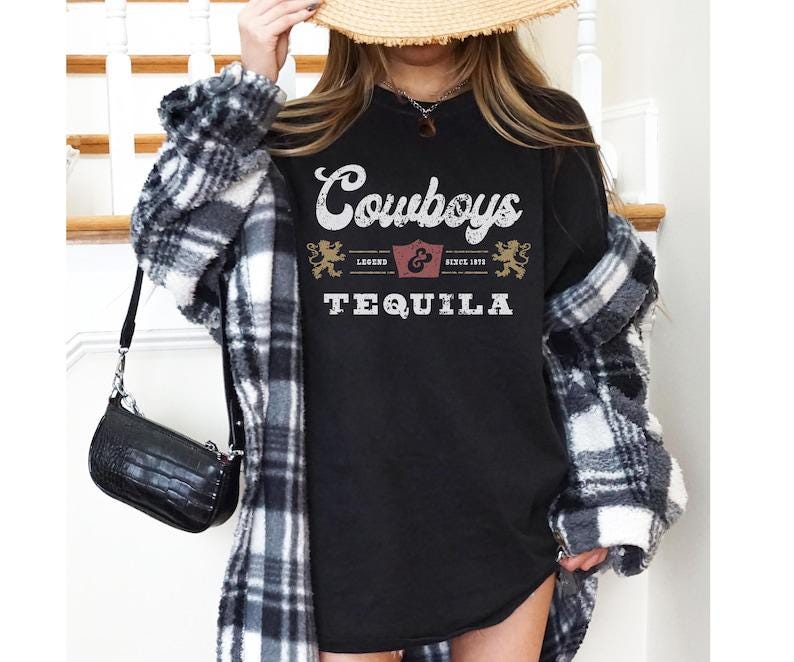 Pu&ograve; includere: T-shirt nero girocollo con le parole "Cowboys & Tequila" in stile vintage. La maglietta ha una grafica a scudo e la scritta "Legend Since 1895". La maglietta &egrave; indossata con una giacca a quadri e una borsa nera.