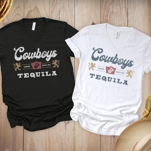 Pu&ograve; includere: Due magliette con scollo a V, una nera e una bianca, con la scritta "Cowboys Tequila" in stile vintage. Il design include uno scudo, due leoni e le parole "Legend" e "Since 1972". Le magliette sono esposte su una superficie di legno.