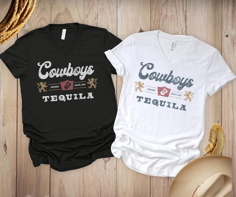 Pu&ograve; includere: Due magliette con scollo a V, una nera e una bianca, con la scritta "Cowboys Tequila" in stile vintage. Il design include uno scudo, due leoni e le parole "Legend" e "Since 1972". Le magliette sono esposte su una superficie di legno.