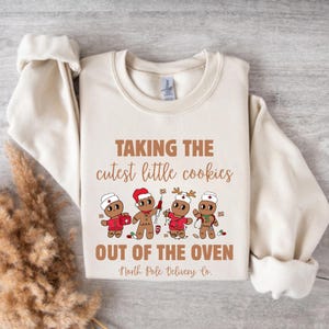 May include: Cream-colored sweatshirt with the text "TAKING THE cutest little cookies OUT OF THE OVEN" in brown. Below the text are four gingerbread cookie characters in red and white outfits. The sweatshirt is a cozy, casual style.