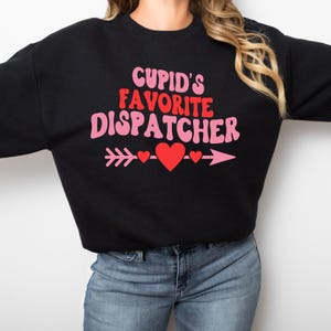 以下が含まれることがあります： ピンクと赤で「CUPID'S FAVORITE DISPATCHER」と書かれた黒いスウェットシャツ。テキストの下には、ハートが付いたピンクの矢印があります。スウェットシャツはクルーネックです。