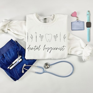 May include: A white crewneck sweatshirt with the words "dental hygienist" in black script, along with dental-themed line art. The image includes blue scrubs, a stethoscope, a name badge, and a light blue smartwatch.