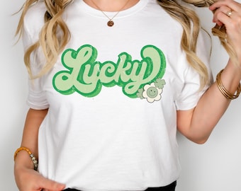 May include: White t-shirt with a green retro-style graphic that says "Lucky" with a green flower and shamrock design.
