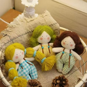 May include: Three handmade dolls with yarn hair in a woven basket. The dolls have rosy cheeks and wear checkered overalls in blue, yellow, and green. The basket also contains pinecones and sits on a beige fabric.