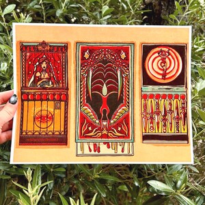 May include: Three vintage-style matchbox labels with illustrations. The first label features a woman in a red dress and a red curtain behind her. The second label features a red and green illustration of a creature with fangs. The third label features a red and yellow illustration of a person on a target with knives around the edge.