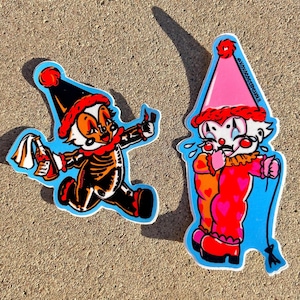 May include: Two clown-themed stickers on a light blue background. One clown is in black and orange, holding a bottle and a rag. The other clown is in pink and red, with tears and a whip. The text "@xoxogossipghoul" is on the pink clown sticker.