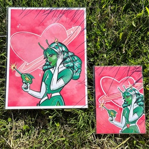 May include: Two framed prints of a green alien woman with a heart-shaped planet behind her. She is holding a ray gun in one hand and has a ring around her finger. The background is pink with white stars.