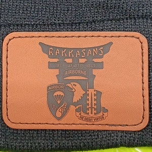 May include: A black knitted beanie with a brown leather patch that reads "RAKKASANS 1ST CAVALRY DIV. AIRBORNE AIRBORNE NO DEBIT VIRTUS"