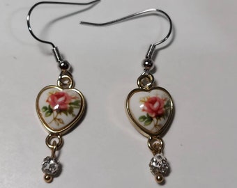 May include: A pair of gold heart-shaped earrings with a white background and a pink rose design. Each earring has a small silver and clear crystal bead dangling from the bottom.