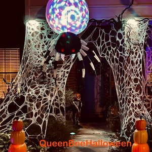 May include: A large inflatable black and white spider with red eyes is hanging from a white spider web. The spider is illuminated by a colorful disco ball. The text "Queen Bee Halloween" is visible in the bottom of the image.
