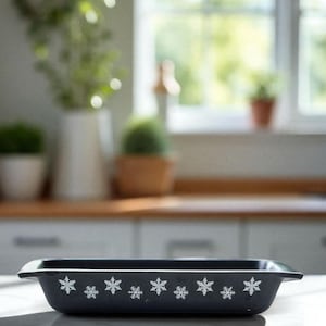 May include: A rectangular, dark blue baking dish with white snowflake designs. The dish has a slightly raised rim and is sitting on a white countertop. The background is a blurred kitchen scene with a window.