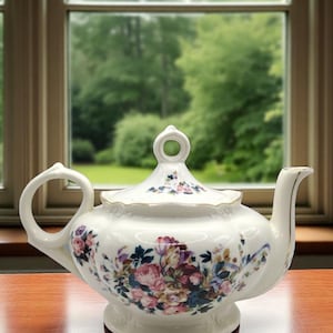 May include: White porcelain teapot with a floral design in pink, purple, and blue. The teapot has a curved spout, a loop handle, and a lid with a small knob. The teapot sits on a wooden surface.