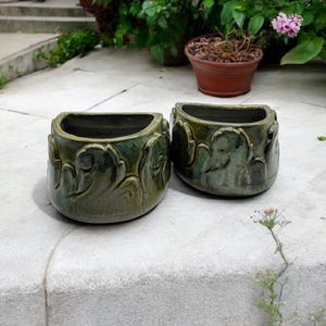 May include: Two green ceramic planters with a decorative leaf design. The planters are half-round and have a glazed finish.