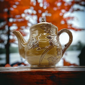 May include: A brown ceramic teapot with an oak leaf design. The teapot has a rounded body and a curved spout. The handle is attached to the side of the teapot.