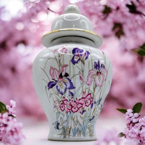May include: A white ceramic jar with a lid, adorned with a floral design featuring purple and pink irises. The jar has a gold trim around the lid and a small knob on top. The background is a soft pink, creating a delicate aesthetic.