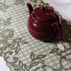May include: A dark red teapot with a lid and a handle. The teapot is sitting on a white tablecloth with a green lace pattern.