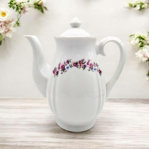 May include: White porcelain teapot with a floral design in shades of purple, pink, and blue. The teapot has a rounded body and a lid with a knob.