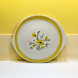 May include: A white ceramic serving platter with a yellow rim and a floral design featuring yellow flowers and brown stems.