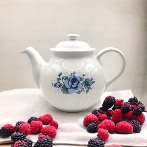 May include: White teapot with a blue floral design and a lid. The teapot is sitting on a white cloth with red and black berries scattered around it.