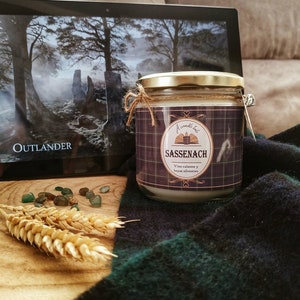 Könnte beinhalten: Ein Kerzengefäß mit einem schwarz-weiß karierten Etikett, auf dem "A candle tale, Sassenach, Vino caliente y bayas silvestres" steht. Die Kerze steht auf einer braunen Oberfläche mit einer grünen Decke und einem Laptop-Bildschirm, auf dem im Hintergrund die Worte "Outlander" zu sehen sind.