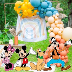 Pode incluir: Um cenário de festa com uma faixa de tecido branco com estampa de arco-íris pastel, cercada por um grande arco de balões em amarelo, azul e pêssego. Figuras recortadas da Minnie, Mickey, Pluto e Pateta estão em primeiro plano.