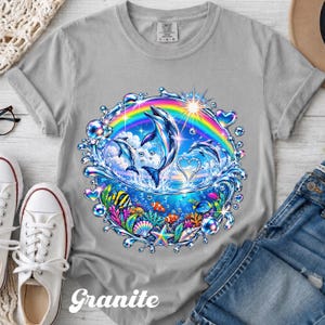 May include: A light gray t-shirt featuring a circular graphic of dolphins leaping from ocean waves under a rainbow. The design includes colorful tropical fish, coral reefs, seashells, and floating bubbles, with a bright sunburst effect.