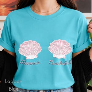 May include: A bright turquoise crew neck t-shirt featuring two light pink seashell illustrations centred on the chest. Below each shell, the text reads Mermaid and Beachclub in a cursive font. The shirt has short sleeves and a classic fit.