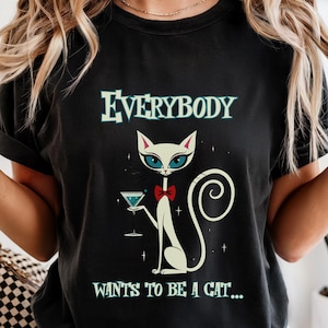 May include: Black t-shirt with the text "EVERYBODY WANTS TO BE A CAT...". The graphic features a white cat with blue eyes, a red bow tie, and a martini glass. The cat has a swirled tail.