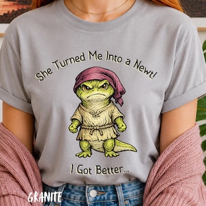 May include: A light gray t-shirt featuring a graphic of a green newt wearing a pink headscarf and a beige tunic. The text above the newt reads She Turned Me Into a Newt! and the text below the newt reads I Got Better... in a dark font.