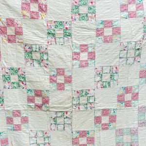 May include: A handmade quilt with a patchwork design. The quilt features alternating squares of white fabric and squares with floral and patterned fabrics in shades of pink and green. The quilt has a vintage, homespun aesthetic.