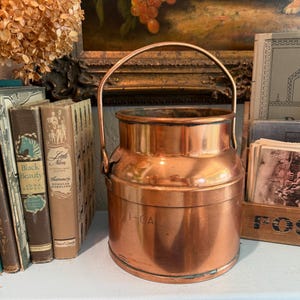 May include: A cylindrical copper milk can with a sturdy arched handle and a wide opening. The exterior surface features a polished metallic finish with the text 1-GAL clearly engraved on the side, indicating its capacity in imperial units.