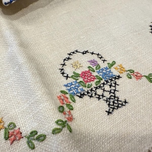 May include: A white linen tablecloth with embroidered floral designs. The embroidery features baskets of flowers in shades of blue, pink, yellow, and green.