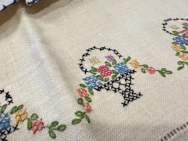 May include: A white linen tablecloth with embroidered floral designs. The embroidery features baskets of flowers in shades of blue, pink, yellow, and green.